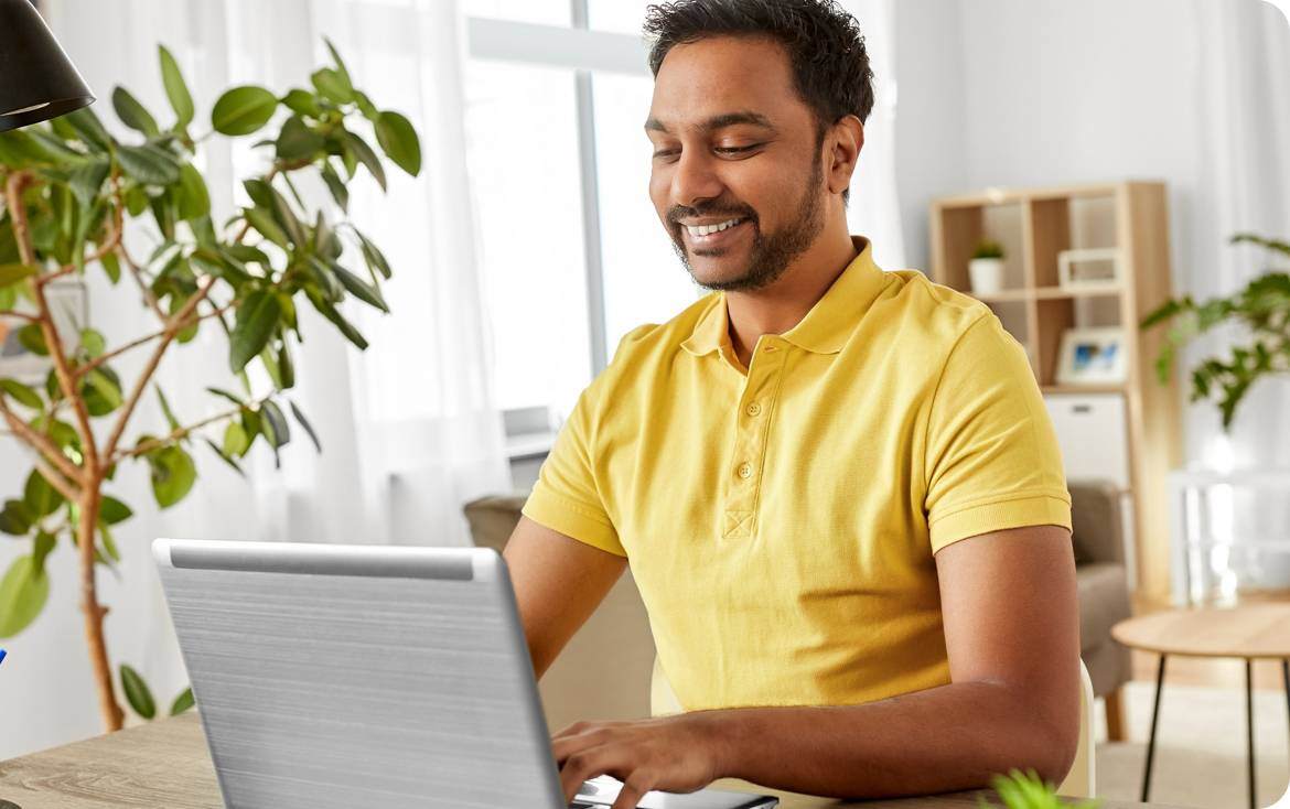 Man wearing a yellow shirt smiling and looking on his laptop in his living room