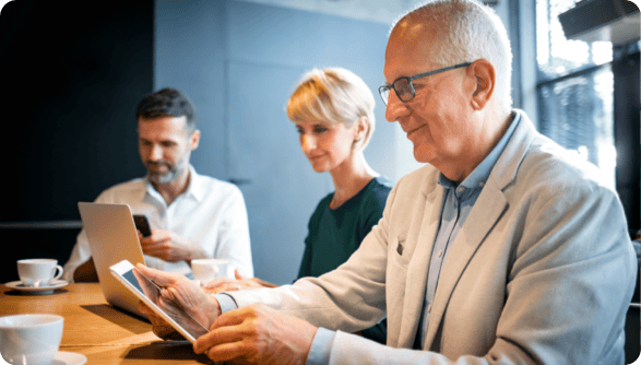 An image of two men and one woman at work, each with a device. The Older man has a tablet, the woman has a laptop and the man is on his phone. There are two mugs of tea or coffee.